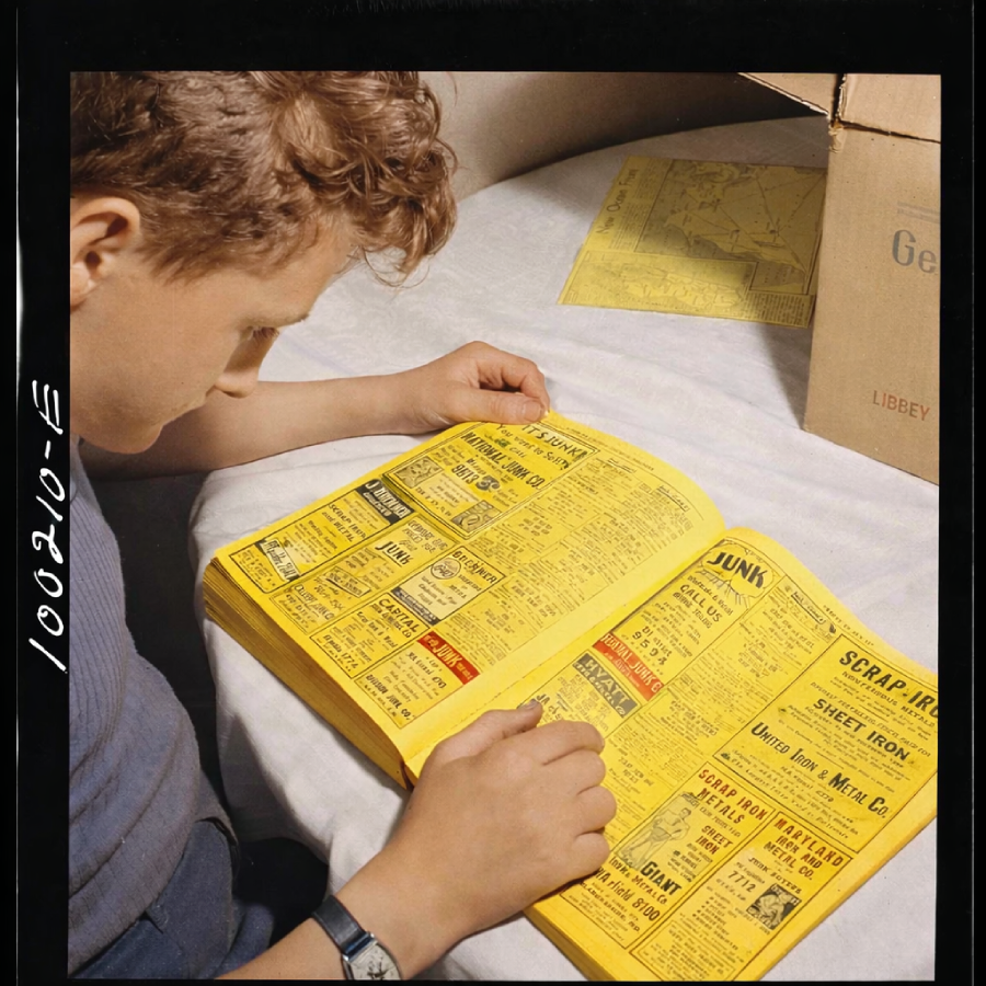 A boy with curly hair sits at a table looking through a yellow phone book, with an open cardboard box and more yellow pages in the background. The table is covered with a white cloth.