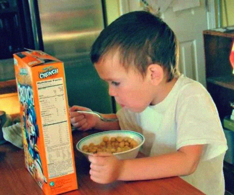 A young boy in a white shirt sits at a kitchen table, eating cereal from a bowl while reading the back of an orange cereal box placed in front of him.