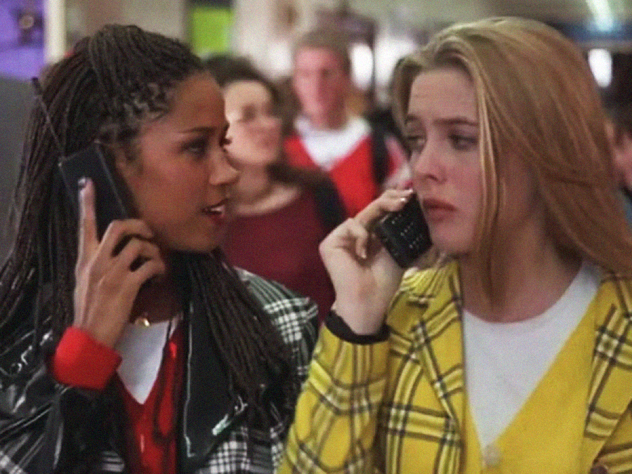 Two young women walk together in a crowded hallway, each holding a cell phone to their ear. One wears a yellow plaid jacket, the other a black-and-white jacket. Both appear to be engaged in conversation.