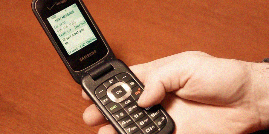 A hand holding a black Samsung flip phone with a green-lit screen displaying a text message over a wooden surface.
