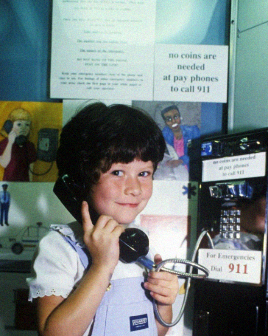 A young girl with short dark hair smiles while holding a payphone receiver to her ear. Behind her are posters, including one stating “no coins are needed at pay phones to call 911.”