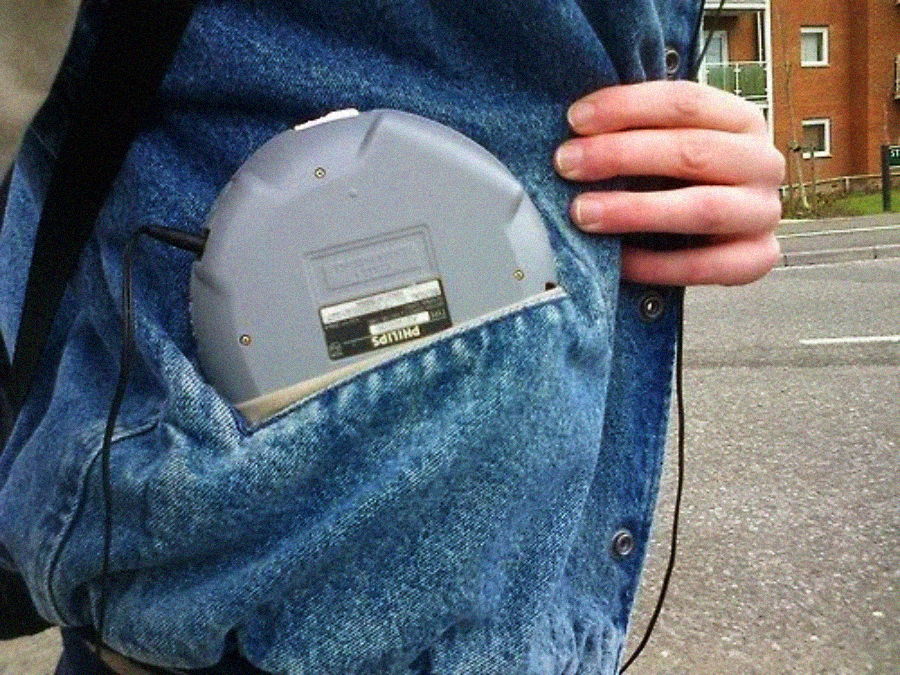 A person wearing a blue denim jacket carries a gray portable CD player partially tucked into their jacket pocket, with headphones plugged in, standing on a street near buildings.