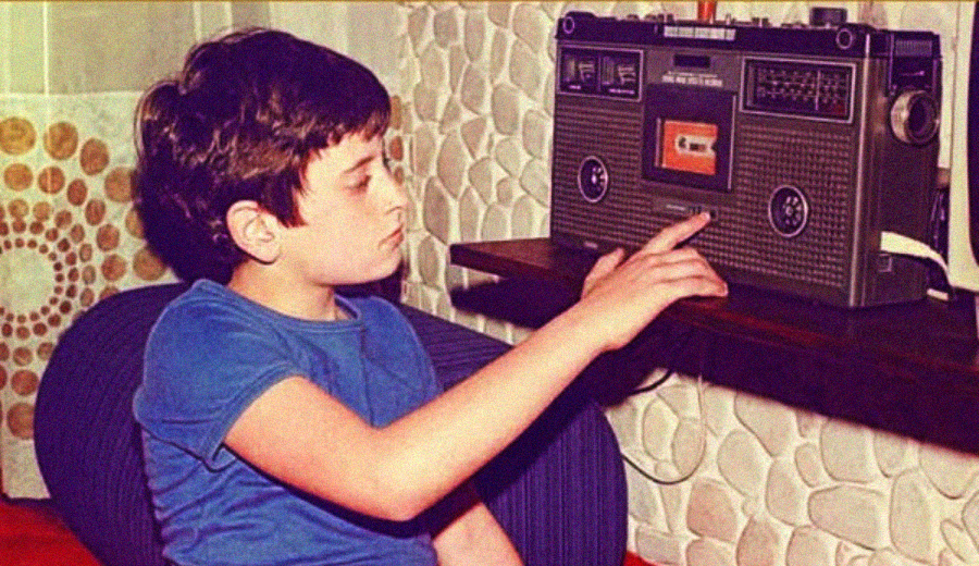 A young boy in a blue shirt sits on a chair, reaching out to press a button on a vintage cassette tape player placed on a table against a patterned wall.
