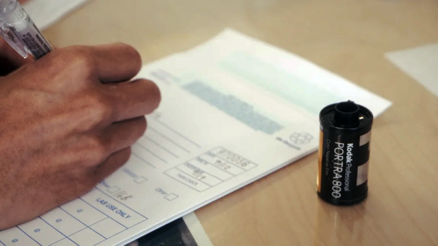 A hand fills out a form with a pen on a desk. Next to the form is a Kodak Professional Portra 800 film canister.