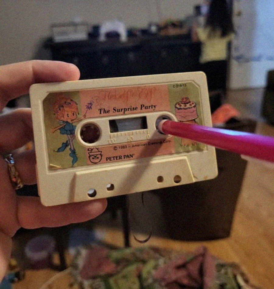 A hand holds a vintage cassette tape labeled "The Surprise Party" with cartoon illustrations, while a pink pencil is inserted into one of the cassette reels. The background shows a room with blurred objects and a person.