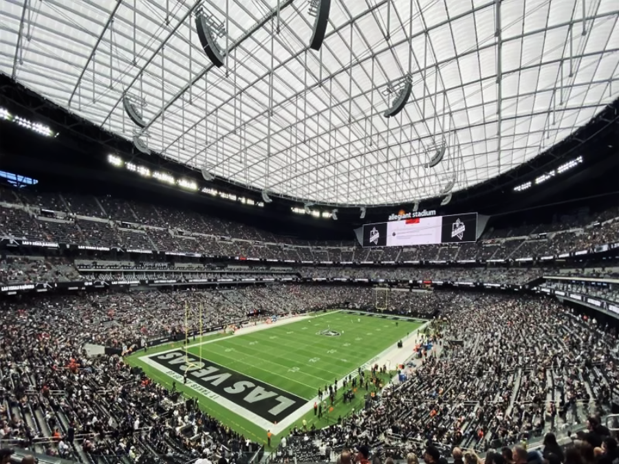 A wide view of a packed football stadium with a transparent roof. The field is marked with "LAS VEGAS" in the end zone, and fans fill the stands, watching the game. Large video screens display information above the crowd.