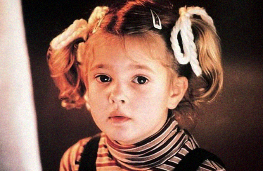 A young child with light brown hair in pigtails tied with white ribbons looks toward the camera. She is wearing a striped turtleneck and dark overalls, with a neutral, slightly serious expression.
