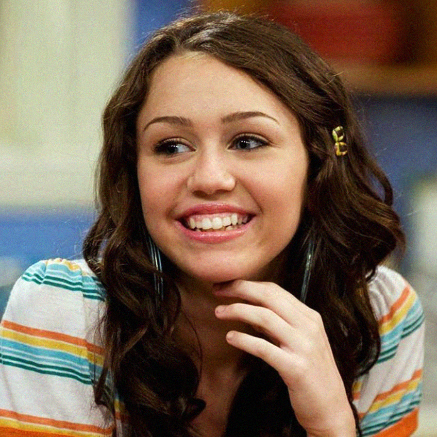 A smiling teenage girl with long, wavy brown hair and a gold hair clip wears a striped shirt and large hoop earrings, resting her hand under her chin. The background is softly blurred.