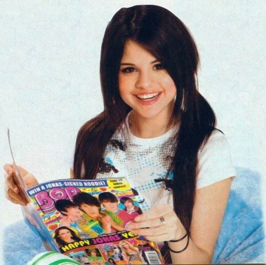 A smiling teenage girl with long dark hair, wearing a white t-shirt, sits holding and reading a colorful teen magazine featuring the Jonas Brothers on the cover.