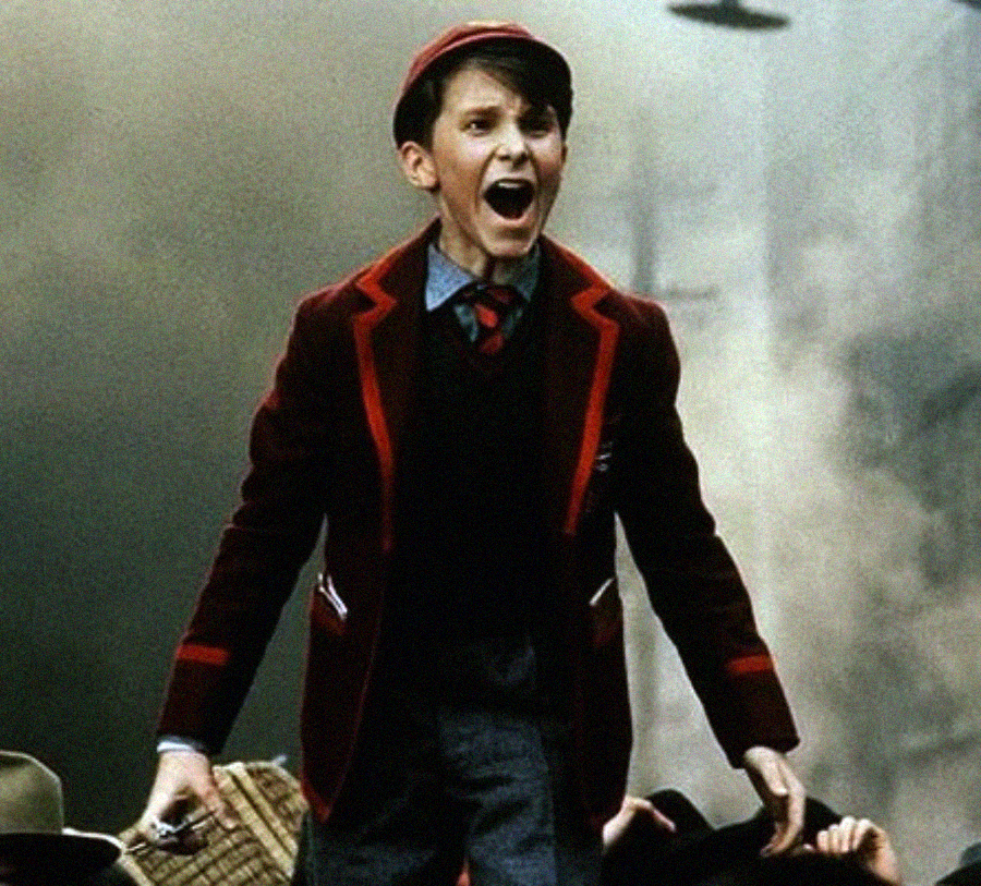A young boy in a red-trimmed school uniform and cap stands outdoors, mouth open as if shouting or singing, with an intense and emotional expression. The background appears smoky and chaotic.