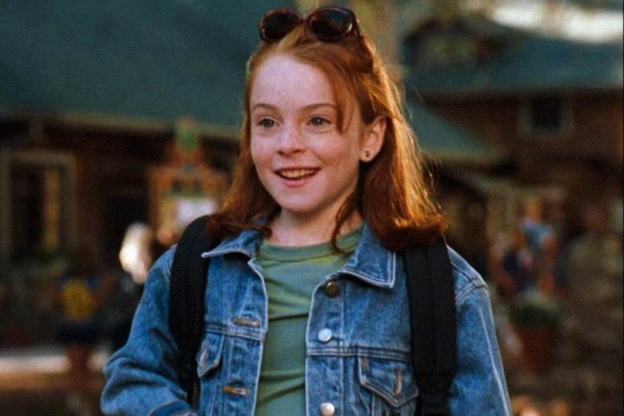 A smiling young girl with red hair wears a denim jacket, green shirt, black backpack, and sunglasses on her head. She stands outdoors with blurred buildings in the background.