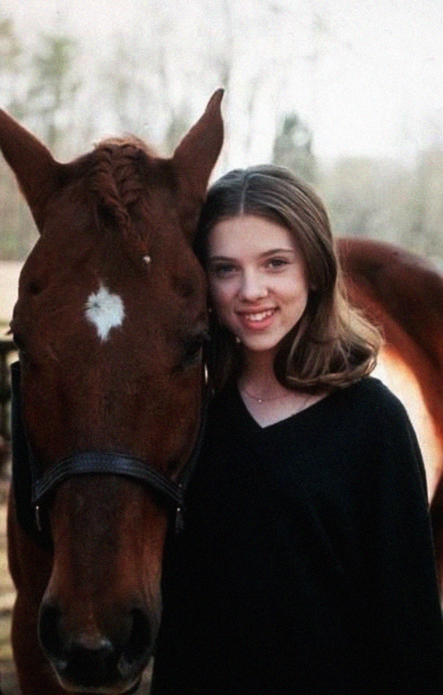 A young woman with shoulder-length brown hair, wearing a black top, stands closely beside a brown horse with a white star marking on its forehead. The pair are outdoors on a lightly blurred background.