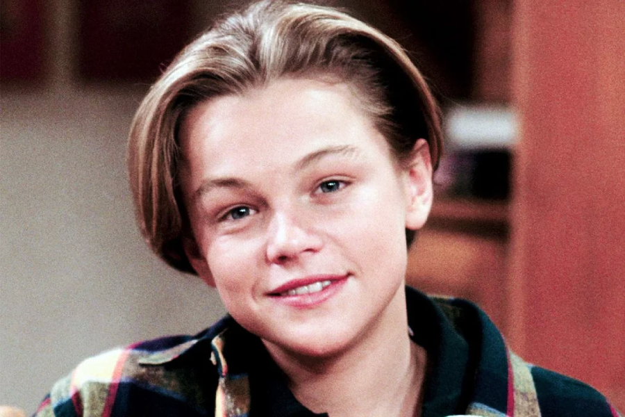 A young person with light skin and short, light brown hair parted in the middle, wearing a plaid shirt and smiling softly while looking at the camera.