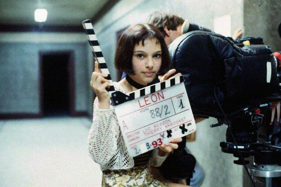 A young girl with short dark hair holds a clapperboard labeled "LEON" in front of a film camera on a movie set, wearing a patterned shirt and standing in a hallway.
