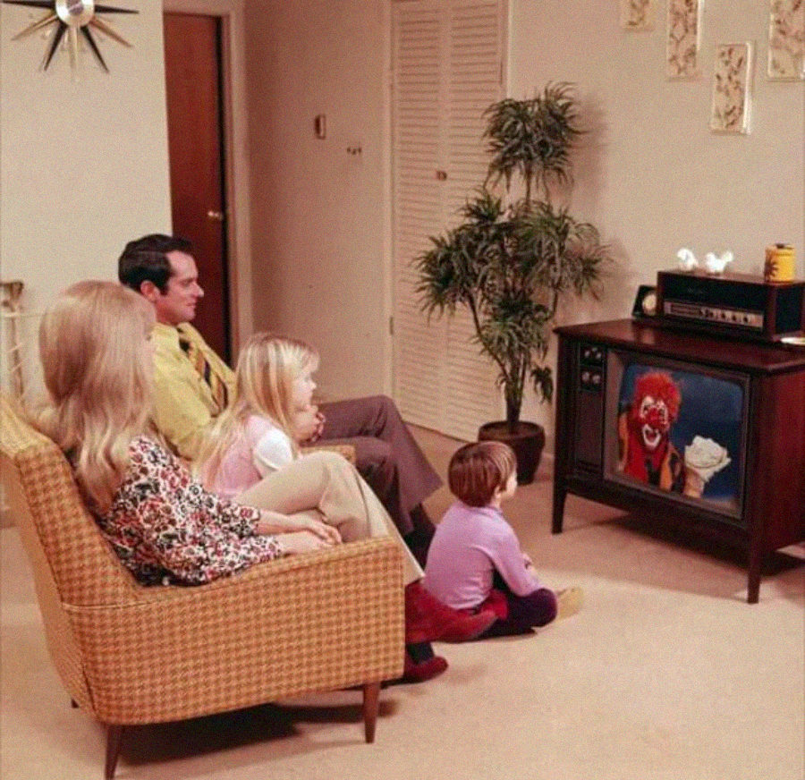 A family of four, including two young children, sits in a retro living room watching a TV showing a clown. The room has tan walls, plants, and vintage decor.