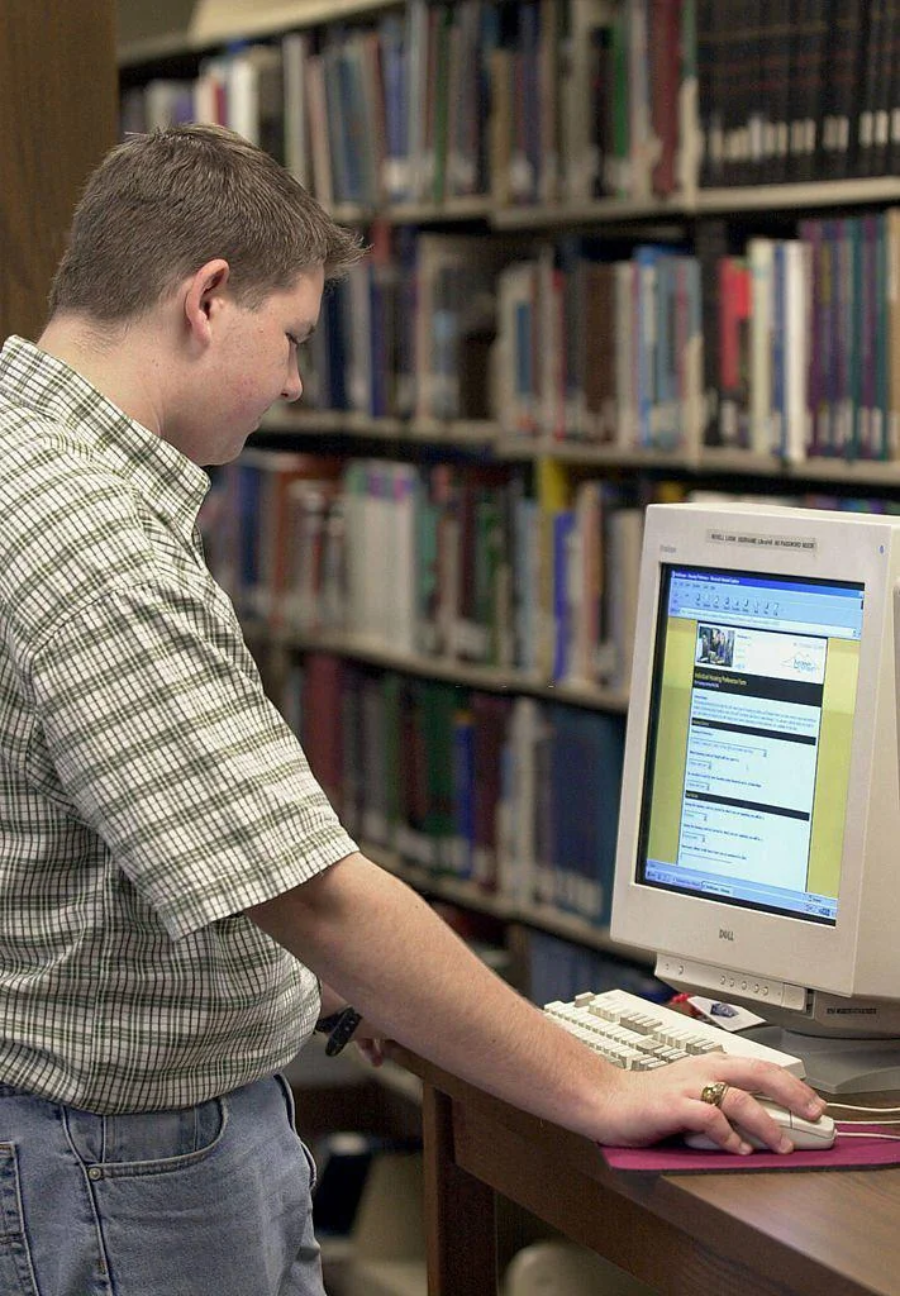 A person stands at a computer terminal in a library, looking at the screen while using the keyboard. Shelves filled with books are visible in the background.