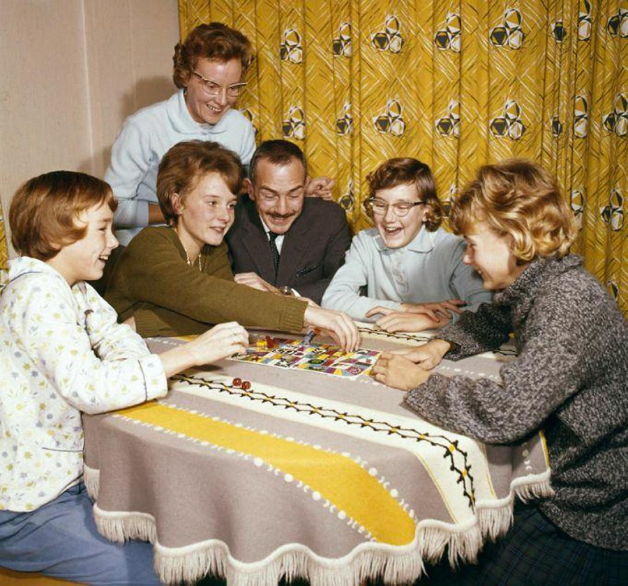 Six people, four women and two men, sit around a table covered with a patterned blanket, smiling and playing a colorful board game in a warmly lit room with yellow patterned curtains in the background.