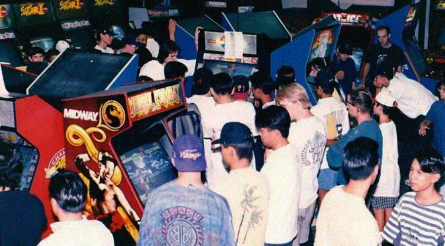 A crowded arcade filled with people playing and watching classic arcade games, including a Mortal Kombat machine in the foreground. The scene is lively and energetic, with rows of game cabinets.