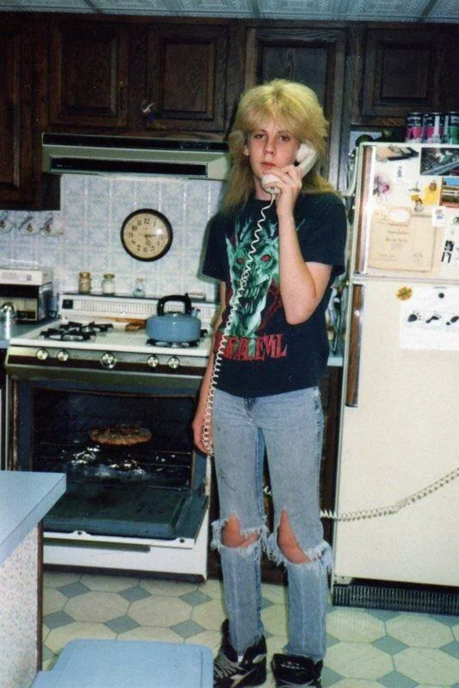 Teen with long blond hair, wearing a graphic t-shirt and ripped jeans, stands in a vintage kitchen talking on a corded phone. Behind them, a pizza is baking in the open oven. The fridge is covered with magnets and photos.