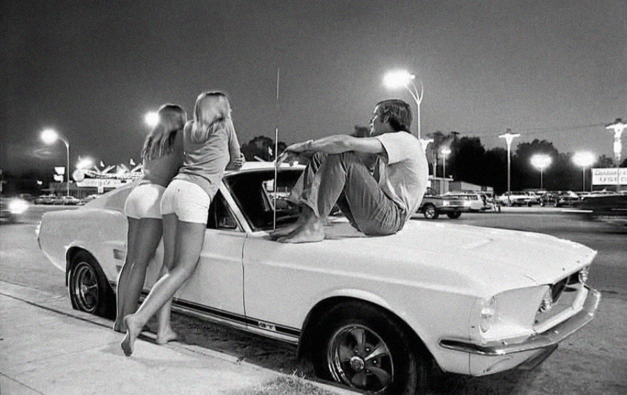 Two women in shorts stand by a classic white car as a man sits barefoot on the hood. It's nighttime, the street is busy, and neon lights from nearby businesses illuminate the scene.