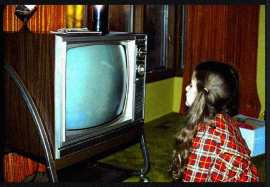 A young girl with long brown hair, wearing a red plaid shirt, sits closely in front of an old-fashioned television with a blank blue screen in a dimly lit room with brown curtains.