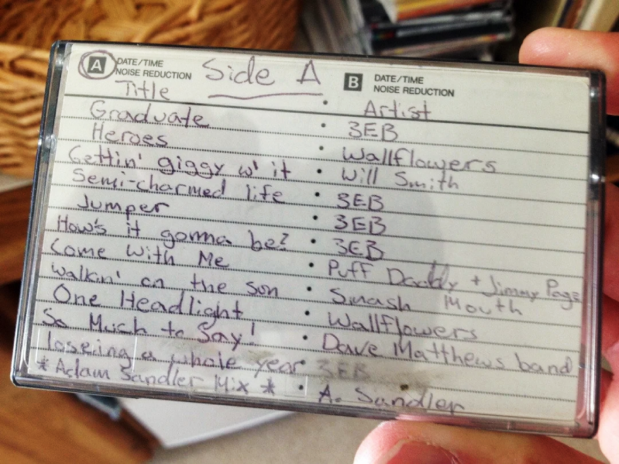 A handwritten mixtape tracklist with the heading "Side A." It lists songs and artists such as "Graduate" by 3EB, "Heroes" by Wallflowers, and "Walkin' on the sun" by Smash Mouth. Fingers hold the case.