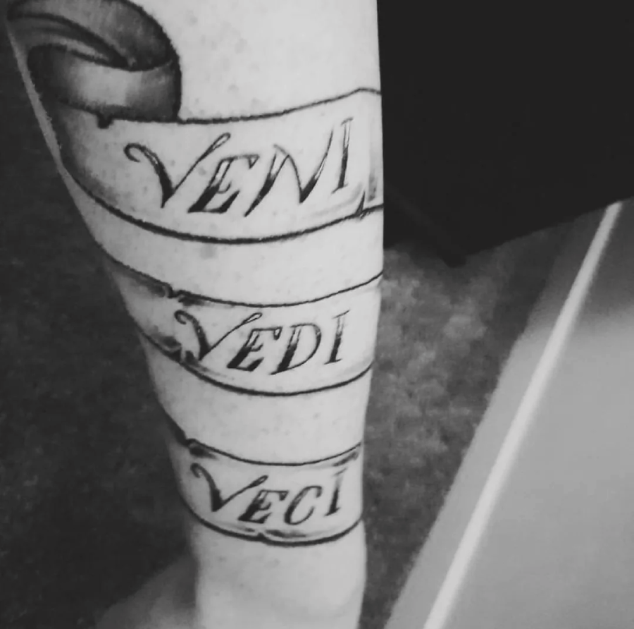 A tattoo on a person's forearm shows a scroll with the Latin words "Veni," "Vidi," and "Vici" written in a script font. The tattoo is in black and gray ink.