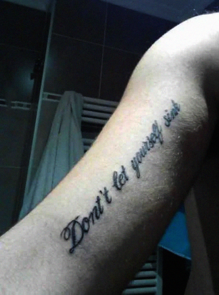 A tattoo on a person's upper arm reads "Don't let yourself sink" in cursive script. The background shows a towel hanging on a hook and tiled walls, suggesting a bathroom setting.