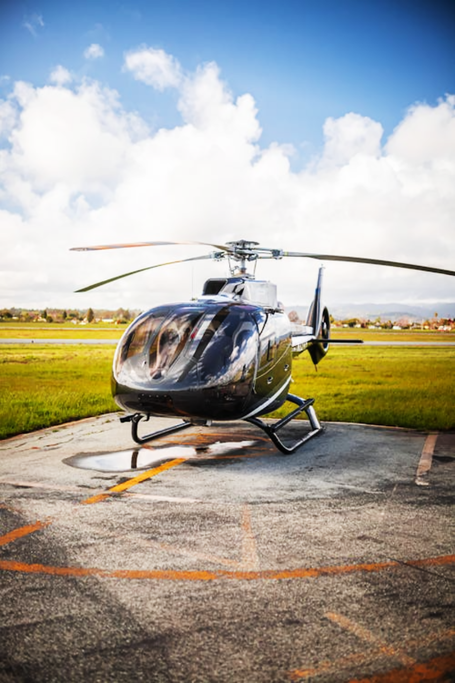 A sleek black helicopter is parked on a helipad with grass fields and a cloudy blue sky in the background.
