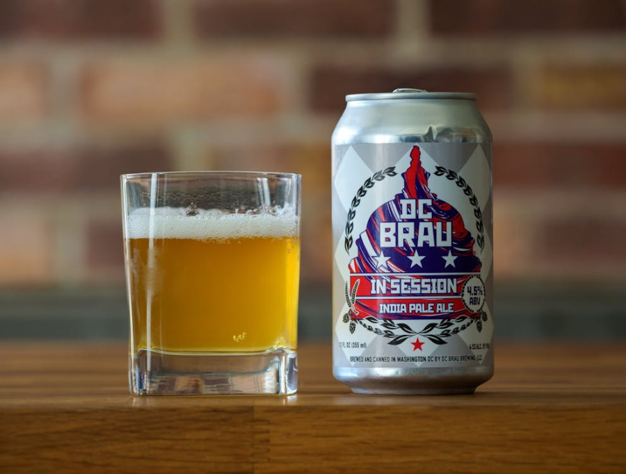 A can of DC Brau In Session India Pale Ale stands next to a glass filled with light amber beer on a wooden surface, with a blurred brick wall in the background.