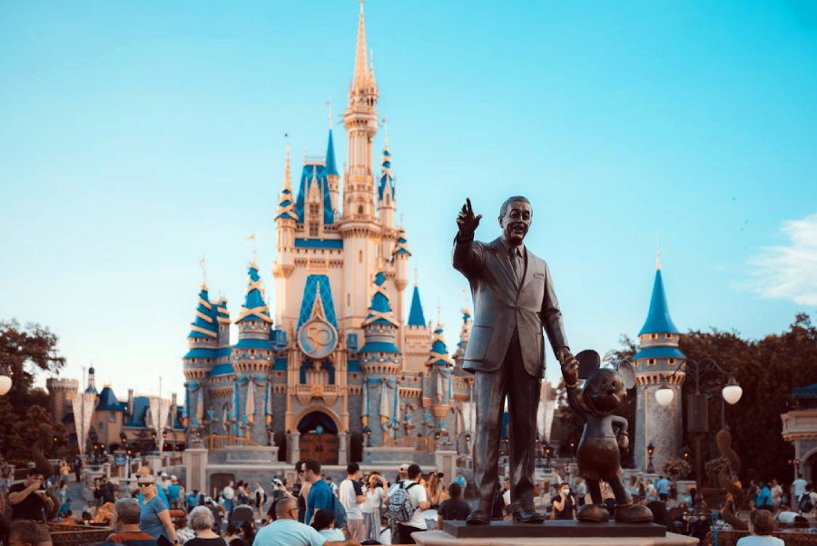 A crowd gathers in front of the Cinderella Castle at Disney World. In the foreground, a statue of Walt Disney holding hands with Mickey Mouse is visible under a clear blue sky.