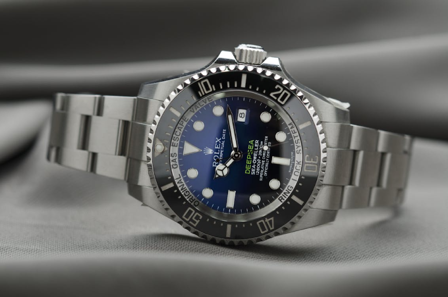 A close-up of a Rolex Deepsea wristwatch with a blue and black gradient dial, silver hour markers, and a stainless steel band, displayed on grey fabric.