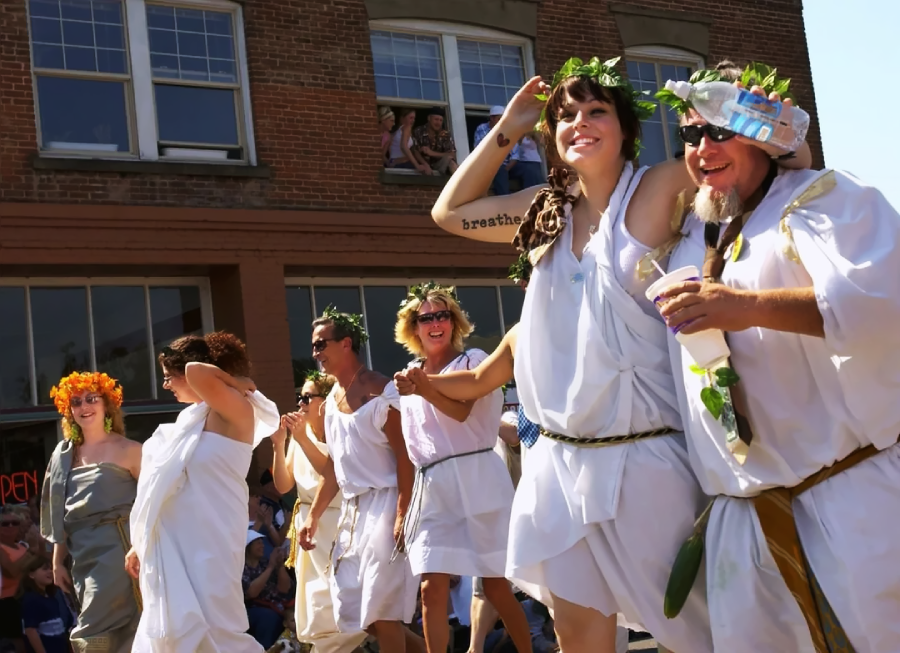 A group of people in white togas and leaf crowns smile and walk in a parade on a sunny day, with brick buildings and onlookers in the background.