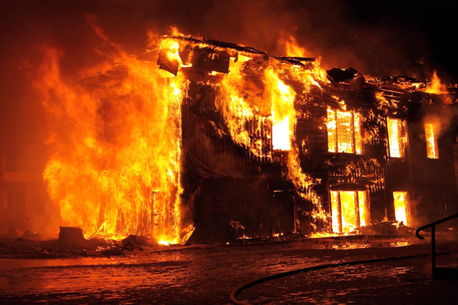 A large building engulfed in bright, intense flames at night, with fire consuming the entire structure and smoke rising into the dark sky. The windows and roof are barely visible through the blaze.