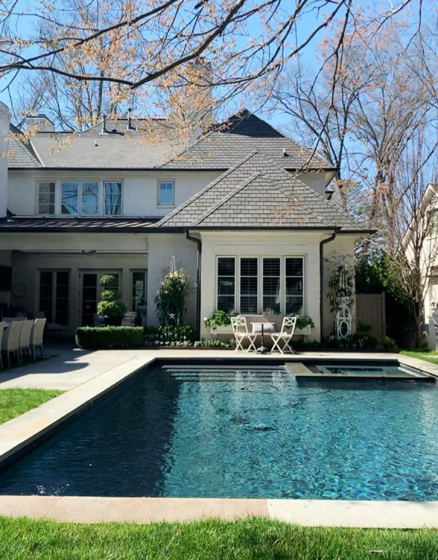 A backyard with a rectangular swimming pool, a grassy lawn, and a patio area featuring a dining table and chairs. The house in the background has large windows and light-colored siding, framed by leafless trees.
