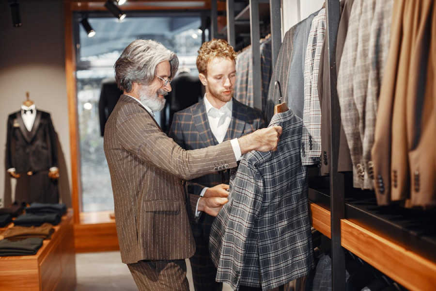 Two men in suits stand in a clothing store, examining a plaid jacket on a rack. The store has wooden shelves and is filled with various suit jackets and formalwear.