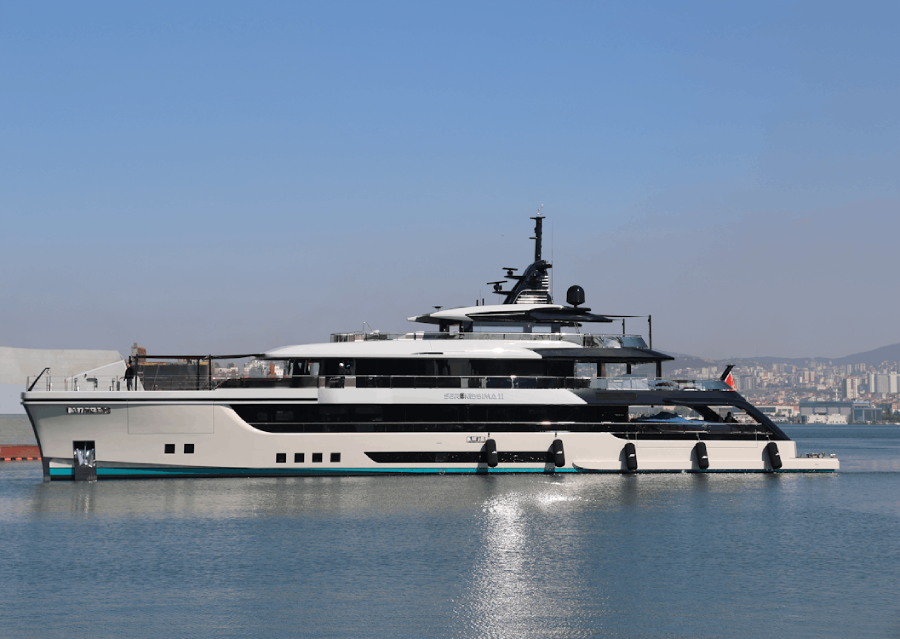 A large, modern white and black yacht is docked in calm water near a city shoreline, with buildings and a clear blue sky in the background.