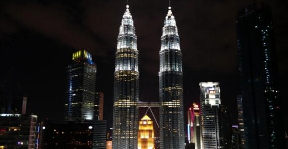 Night view of Kuala Lumpur&rsquo;s skyline featuring the illuminated Petronas Twin Towers at the center, surrounded by other high-rise buildings with city lights against a dark sky.