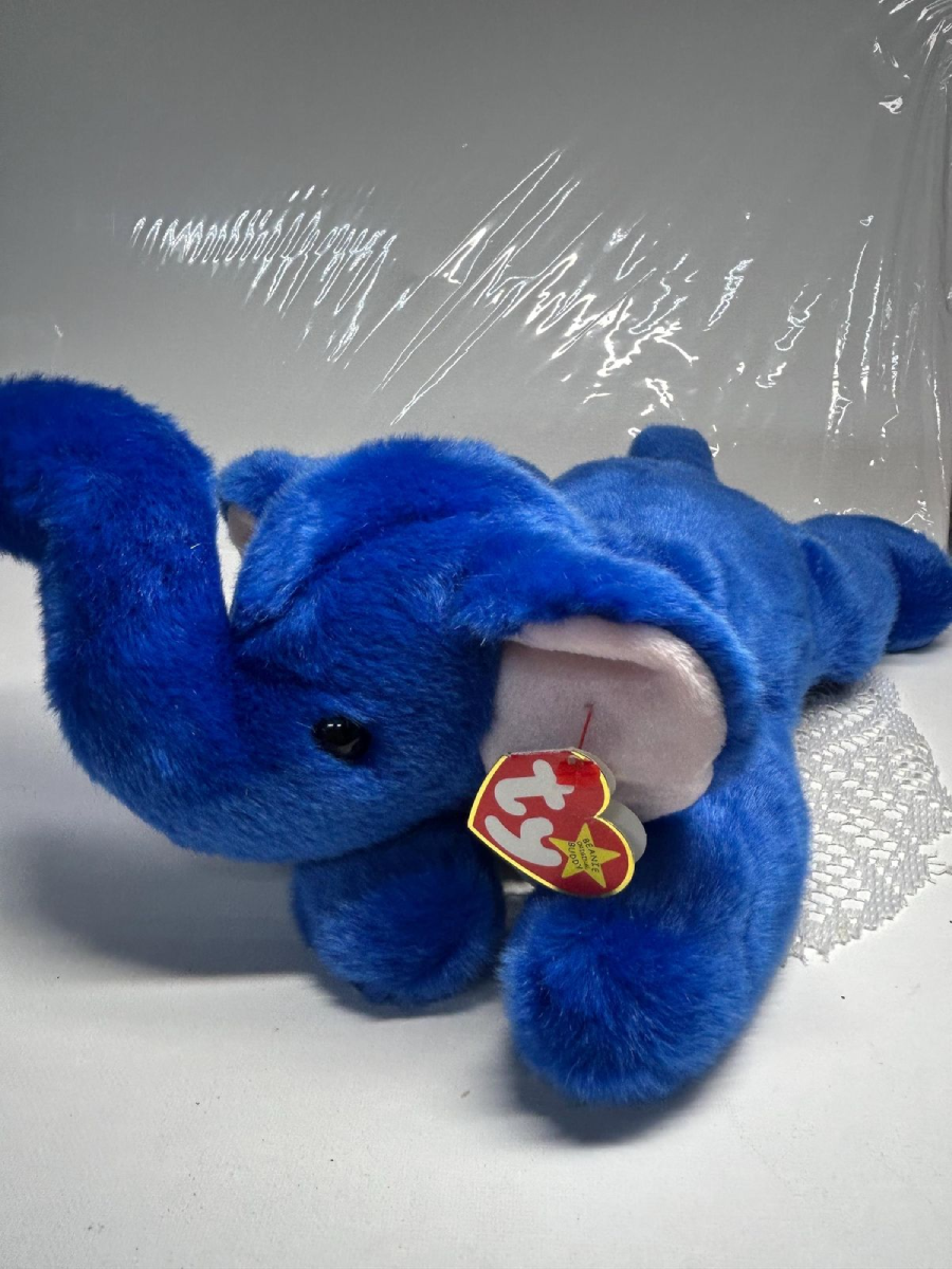 A blue plush elephant Beanie Baby with pink inner ears and a TY tag attached, lying on a white surface with a clear plastic backdrop.