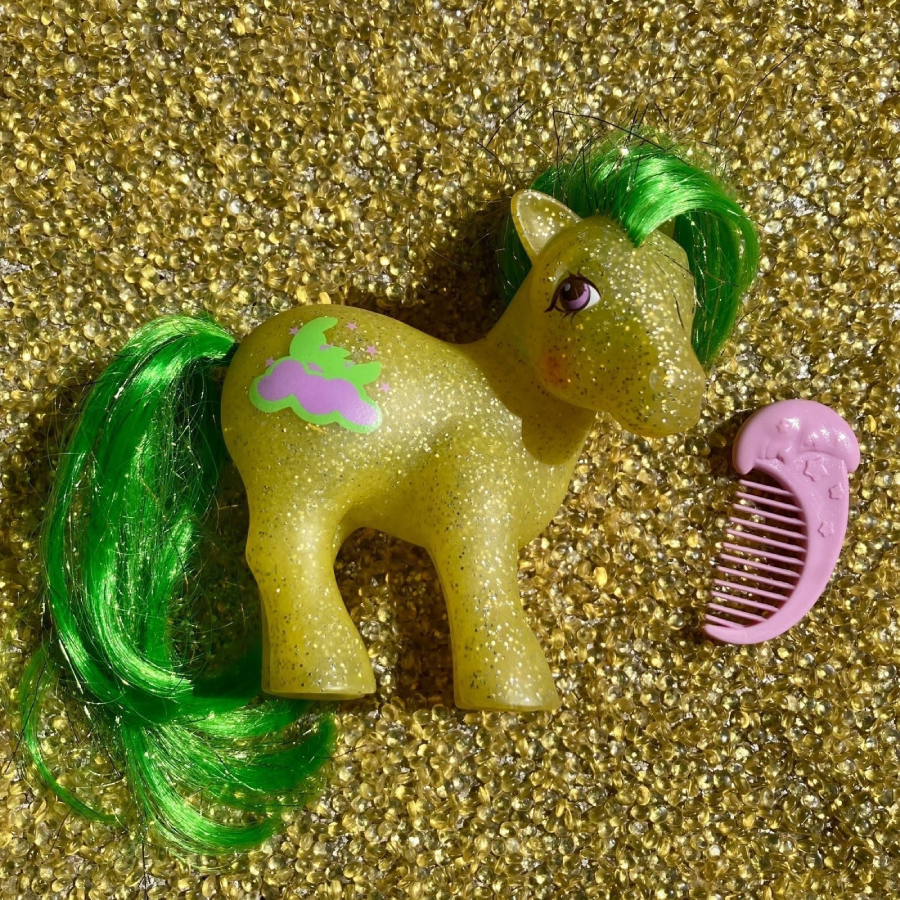 A glittery yellow toy pony with green hair, a pink comb, and a green bunny design on its side, rests on a sparkly gold background.