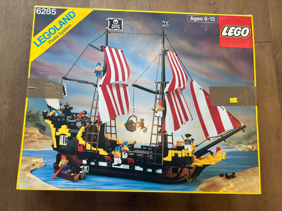 A box of LEGO set 6285, featuring a pirate ship with red-and-white striped sails, minifigures, and accessories. The box shows the ship on water with a sunset tropical background. For ages 9-12.