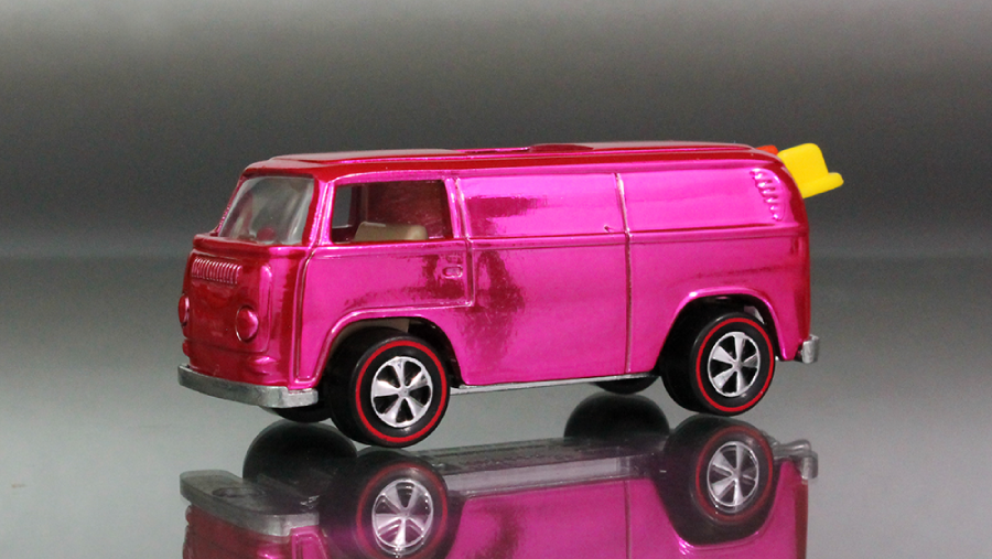 A shiny pink toy van with black and silver wheels is shown on a reflective surface. The van has a yellow surfboard attached to the roof at the back.