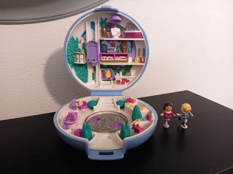 A round, open Polly Pocket toy case with detailed miniature rooms and accessories is displayed on a table. Two small doll figures stand beside it. The scene includes trees, shelves, and tiny furniture in bright colors.