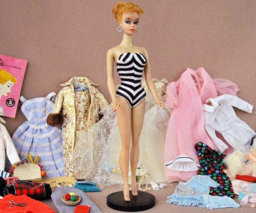 A vintage Barbie doll wearing a black-and-white striped swimsuit stands on a display stand, surrounded by dresses, accessories, and doll clothes laid out on a table.