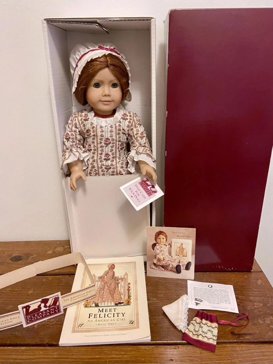 An American Girl doll dressed in colonial-style clothing sits in an open box. The set includes a booklet, a card, a pamphlet, a paper hat, and a small drawstring bag, all displayed on a wooden surface.