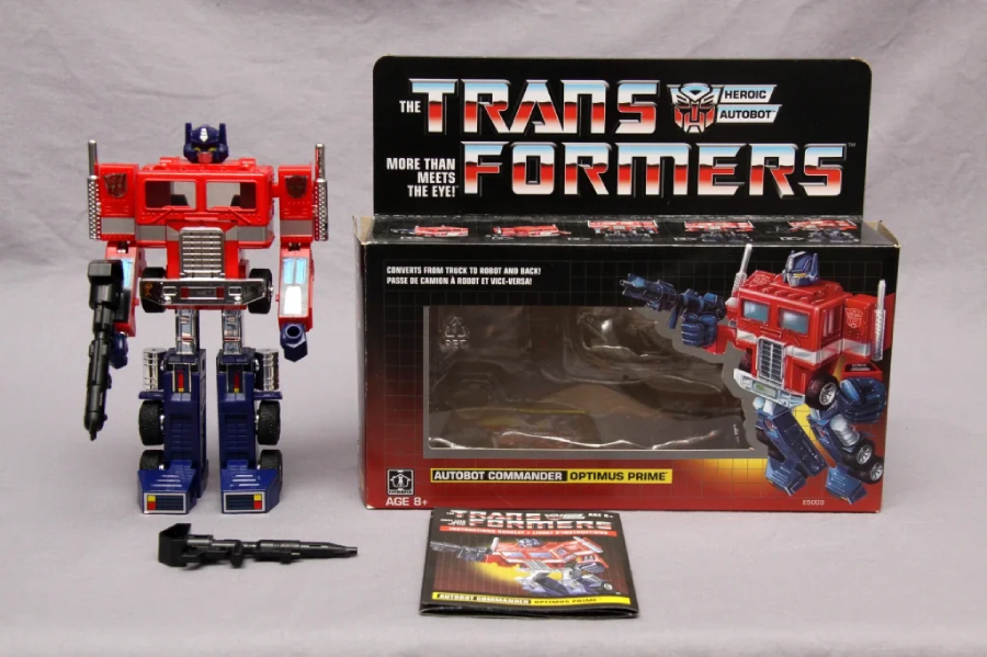 A vintage Transformers Optimus Prime toy stands next to its original box and instruction manual. The box features artwork of Optimus Prime in truck form and the Transformers logo. A black accessory is placed in front.