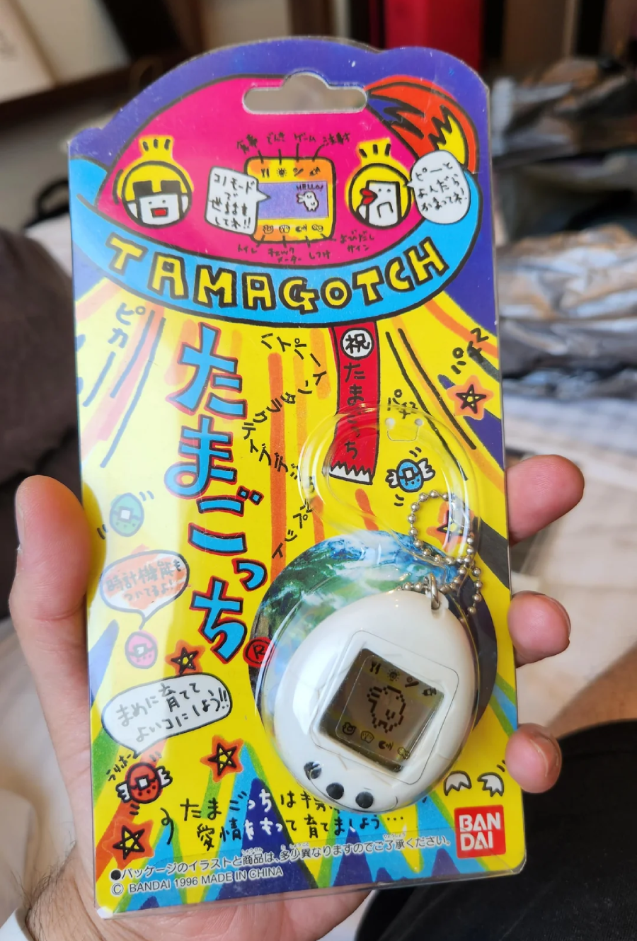 A hand holds a packaged white Tamagotchi keychain toy with colorful Japanese text and graphics on the cardboard backing. The packaging features bright yellow, blue, and red designs.