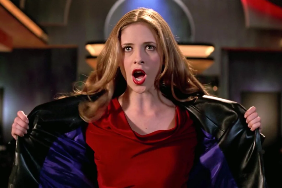 A woman with long hair, wearing a red top and black jacket, stands with her mouth open and holds her jacket open in a dramatic pose indoors.