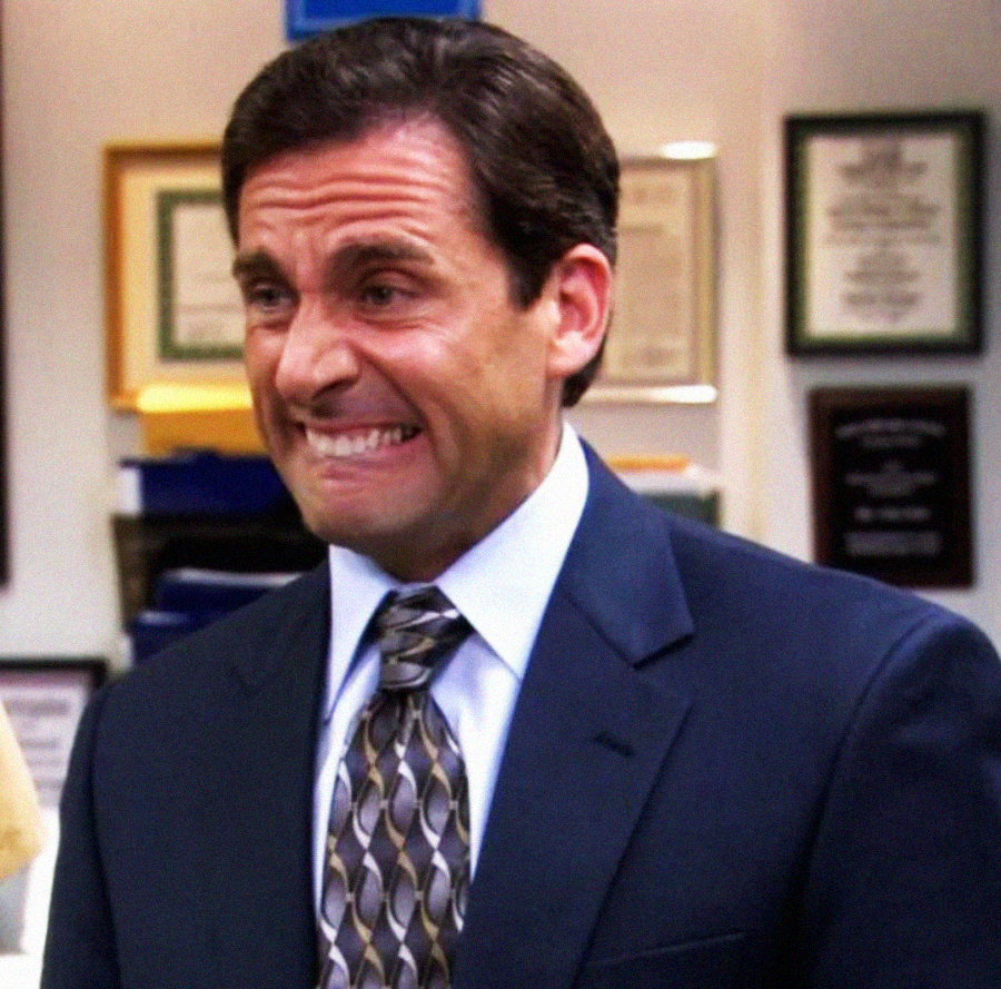A man in a suit with dark hair makes an awkward, uncomfortable grimace while standing in an office with certificates and plaques on the wall behind him.