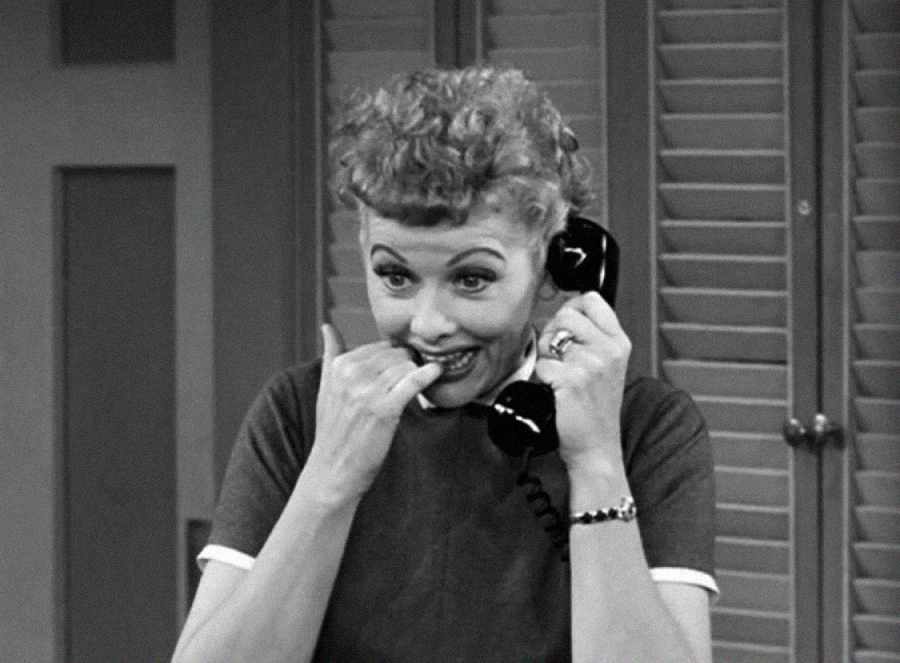 A woman with curly hair smiles while talking on a black rotary phone, holding the receiver to her ear. She playfully bites her finger, appearing amused. The image is in black and white.